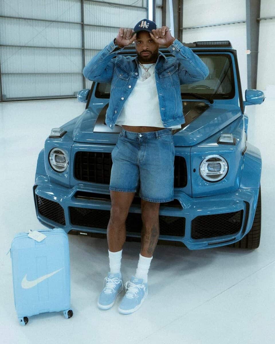 A man in a blue shirt and denim jacket standing next to a blue car.