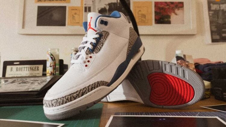 Air Jordan 3 "True Blue" IF4396-104 white leather sneaker with True Blue accents, cement grey elephant print, and red Jumpman detailing, displayed on a wooden desk
